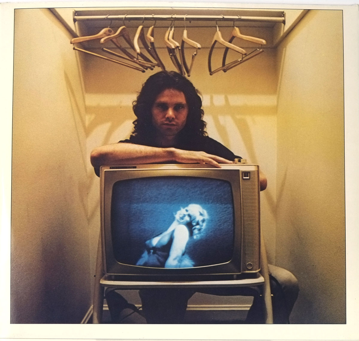 Inner sleeve photo of An American Prayer featuring Jim Morrison seated in a narrow space with empty wooden hangers above him, leaning on a vintage television displaying a glamorous woman, creating a surreal and introspective atmosphere.